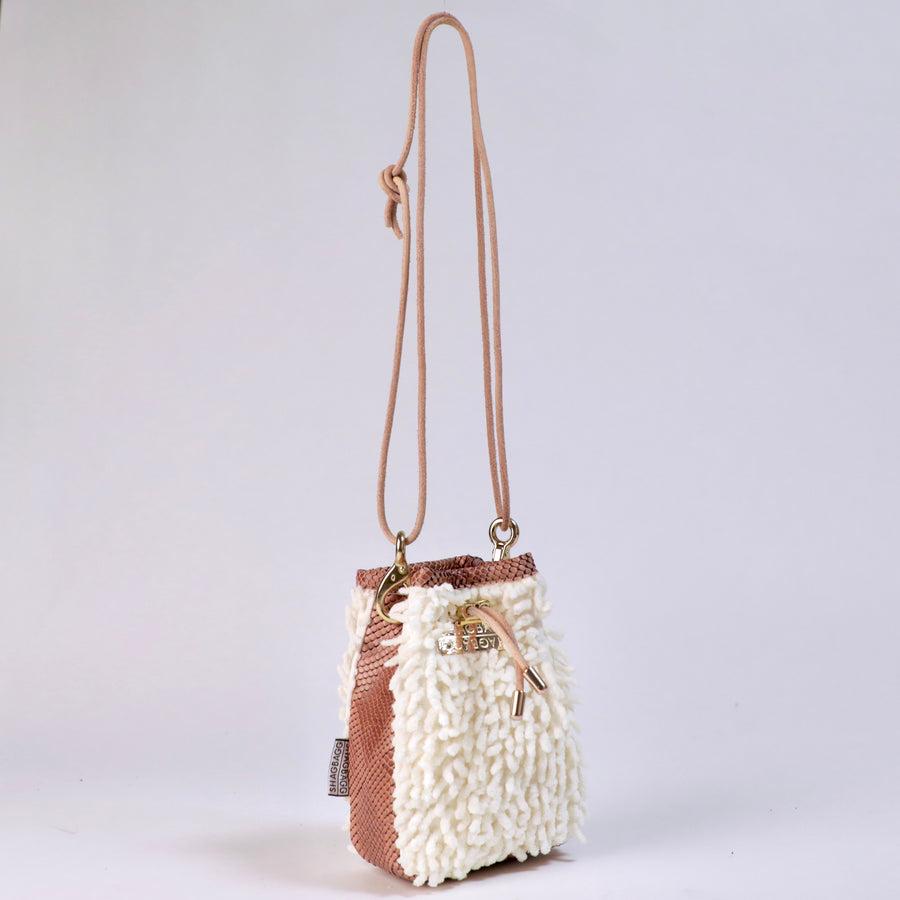 A small bucket-style handbag with a textured white shaggy exterior, a brown snakeskin-patterned side panel, a tan shoulder strap, and gold-tone hardware including a clasp and metal plate that says, SHAGBAGG.
