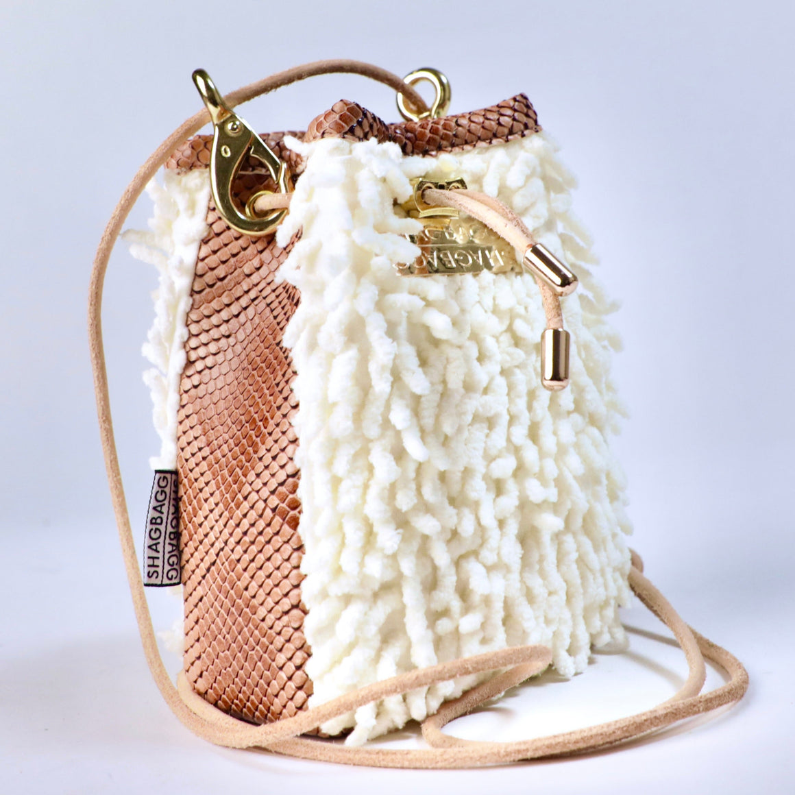 A small bucket-style handbag with a textured white shaggy exterior, a brown snakeskin-patterned side panel, a tan shoulder strap, and gold-tone hardware including a clasp and metal plate that says, SHAGBAGG.
