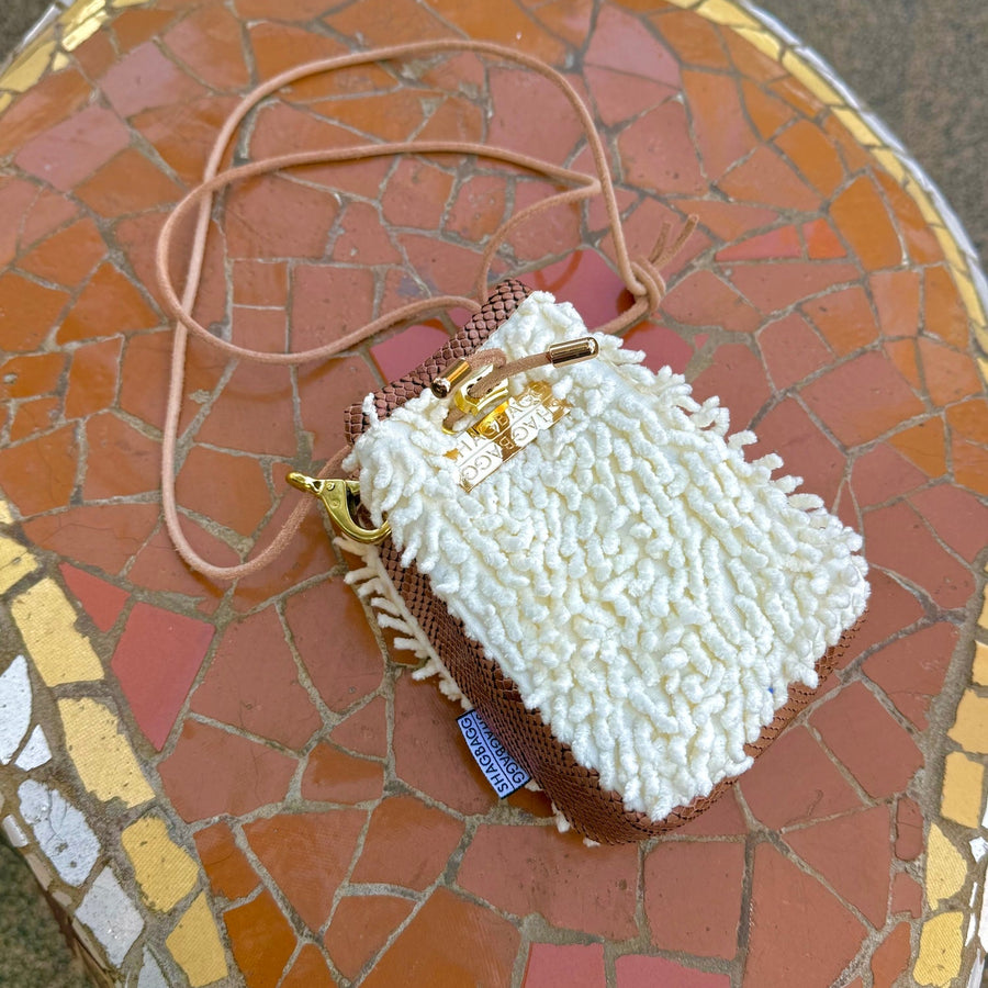 A small bucket-style handbag with a textured white shaggy exterior, a brown snakeskin-patterned side panel, a tan shoulder strap, and gold-tone hardware including a clasp and metal plate that says, SHAGBAGG.